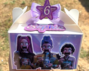May include: A white party favor box with a purple holographic design. The box features three cartoon characters and the name "Sophia". A purple star with the number "6" and a bow adorn the top. The box is held by a hand.