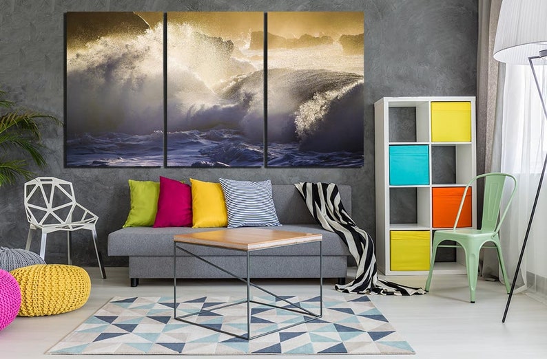 May include: A living room interior design with a gray sofa, a coffee table, a white chair, a green chair, a bookshelf with colorful boxes, and a large canvas print of crashing ocean waves.