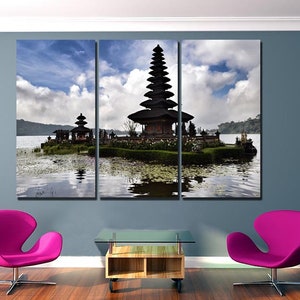 May include: A three-panel canvas print featuring a scenic view of a traditional Balinese temple on a small island in a lake. The temple is a multi-tiered structure with a dark wood finish. The lake is calm and reflective, with a blue sky and white clouds in the background.