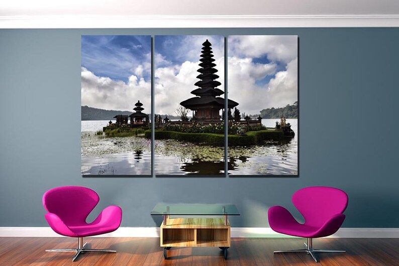 May include: A three-panel canvas print featuring a scenic view of a traditional Balinese temple on a small island in a lake. The temple is a multi-tiered structure with a dark wood finish. The lake is calm and reflective, with a blue sky and white clouds in the background.