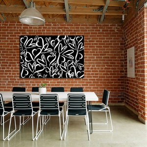 Puede incluir: Una impresi&oacute;n en lienzo en blanco y negro con un dise&ntilde;o de corazones blancos sobre un fondo negro. La impresi&oacute;n est&aacute; colgada en una pared de ladrillo en una habitaci&oacute;n con una mesa blanca y sillas negras.