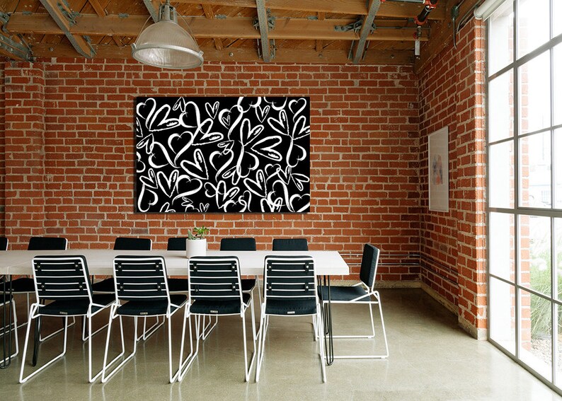 Puede incluir: Una impresi&oacute;n en lienzo en blanco y negro con un dise&ntilde;o de corazones blancos sobre un fondo negro. La impresi&oacute;n est&aacute; colgada en una pared de ladrillo en una habitaci&oacute;n con una mesa blanca y sillas negras.