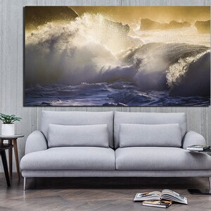 May include: A large canvas print of a dramatic ocean scene with crashing waves and a golden sunset. The waves are white and blue, and the sunset is a warm yellow and orange. The print is hanging on a gray wall above a gray sofa.