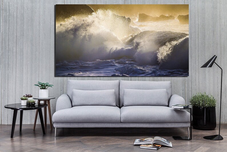 May include: A large canvas print of a dramatic ocean scene with crashing waves and a golden sunset. The waves are white and blue, and the sunset is a warm yellow and orange. The print is hanging on a gray wall above a gray sofa.