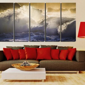 May include: A five-panel canvas print depicting a dramatic seascape with crashing waves and golden sunlight. The print is displayed on a wall above a brown couch with red pillows and a white coffee table.