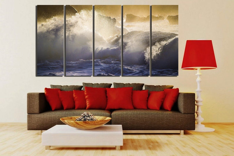 May include: A five-panel canvas print depicting a dramatic seascape with crashing waves and golden sunlight. The print is displayed on a wall above a brown couch with red pillows and a white coffee table.