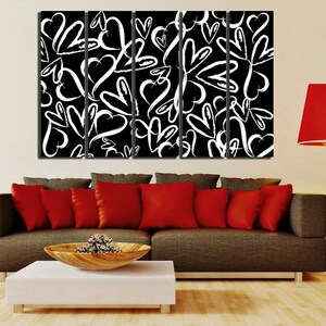 Puede incluir: Una impresi&oacute;n de arte de pared de lienzo en blanco y negro con un patr&oacute;n repetitivo de corazones blancos sobre un fondo negro. La impresi&oacute;n est&aacute; dividida en cinco paneles y se muestra sobre un sof&aacute; marr&oacute;n con almohadas rojas.