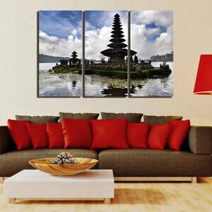 May include: A three-panel canvas print featuring a scenic view of a traditional Balinese temple on a small island in a lake. The temple is dark brown with a tiered roof and is surrounded by lush greenery. The lake is calm and reflective, with a blue sky and white clouds in the background.