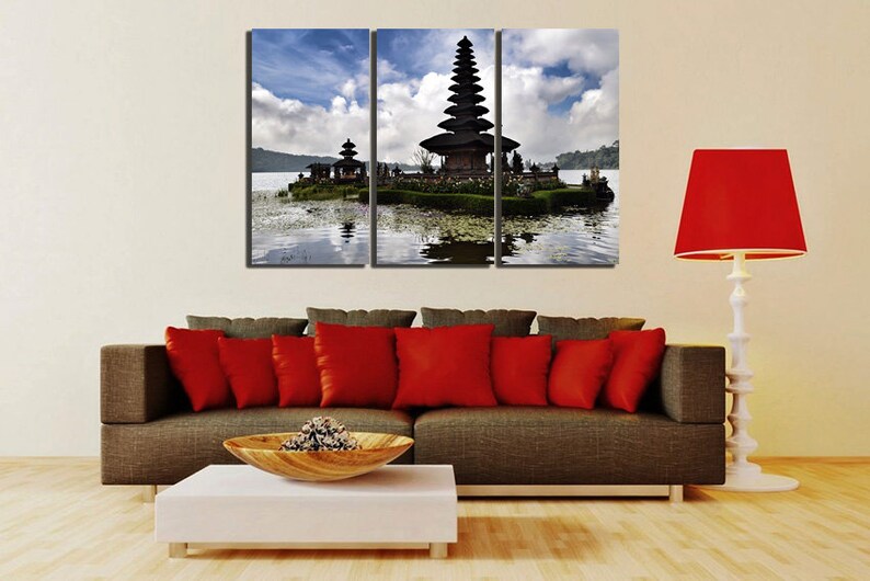 May include: A three-panel canvas print featuring a scenic view of a traditional Balinese temple on a small island in a lake. The temple is dark brown with a tiered roof and is surrounded by lush greenery. The lake is calm and reflective, with a blue sky and white clouds in the background.