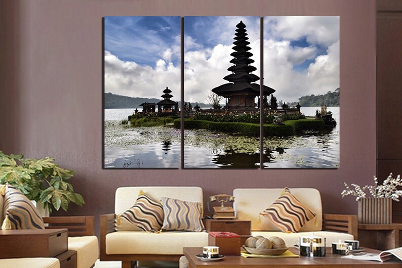 May include: A three-panel canvas print featuring a scenic view of a traditional Balinese temple on a lake. The temple is a multi-tiered structure with a thatched roof, surrounded by lush greenery and water lilies. The sky is a bright blue with fluffy white clouds.