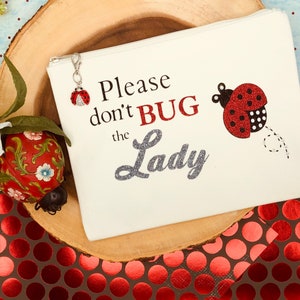 May include: White zippered pouch with the text "Please don't BUG the Lady" in black and silver glitter. A red ladybug with black spots is on the right side of the pouch. A small red ladybug charm is attached to the zipper.