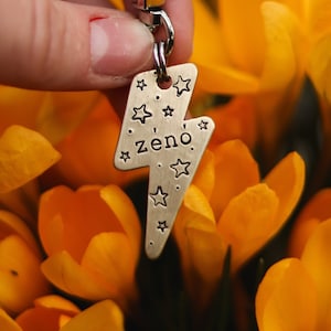 Peut inclure: Breloque en forme d'éclair argenté avec le nom "zeno" gravé dessus, entouré de petites étoiles. Le charme est attaché à un fermoir pivotant argenté et est suspendu à une main.