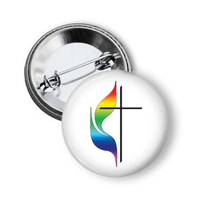 May include: A round pinback button featuring a white background with a black cross symbol overlaid on a stylized flame graphic colored in a rainbow gradient. The back of the button is metallic silver with a standard pin mechanism for attachment.