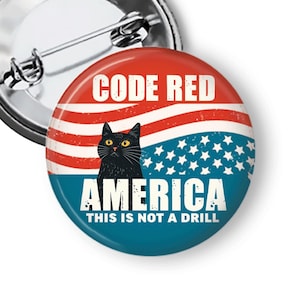 May include: A round button with a red and blue design, featuring a black cat with yellow eyes. The button has the text "CODE RED AMERICA THIS IS NOT A DRILL" in white. A silver pin back is visible.