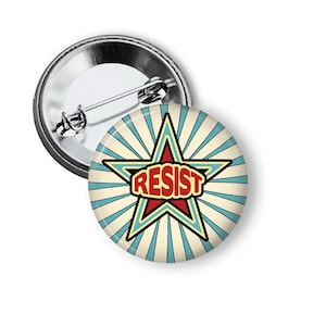 May include: A round button pin with a star design and the word "RESIST" in red. The star is outlined in black and blue, set against a background of blue and cream-colored rays. The pin has a metal backing.