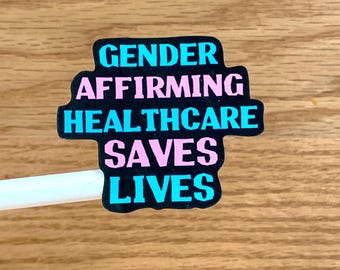 May include: A sticker with the text "GENDER AFFIRMING HEALTHCARE SAVES LIVES" in a black, cross-shaped design. The words are in a combination of light blue and pink, set against a wooden background.