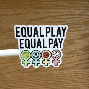 May include: A white sticker with the text "EQUAL PLAY EQUAL PAY" in bold black letters. Below the text are four gender symbols, each with a different sports ball: tennis, soccer, basketball, and baseball.
