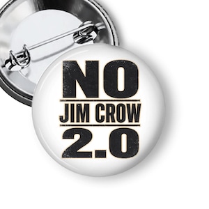 May include: A white button pin with the text "NO JIM CROW 2.0" in black, distressed font. The pin has a metal backing with a safety pin closure.