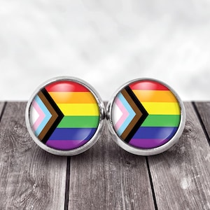 May include: A pair of round stud earrings featuring the Progress Pride flag design. Each earring has a silver-toned metal frame surrounding a circular insert displaying the horizontal rainbow stripes with a black, brown, light blue, pink, and white chevron.