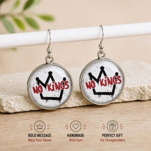 May include: A pair of round silver earrings. Each earring features a white background with a black crown design and the words "NO KINGS" in red. The design has a graffiti-style aesthetic.