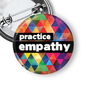 May include: A round button with a pin back. The button has a colorful geometric design in shades of red, orange, yellow, green, blue, and purple. The words "practice empathy" are printed in white on a black band.