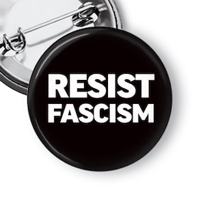 May include: A circular pin-back button featuring a solid black background with the text RESIST FASCISM printed in bold white capital letters. The metallic pin mechanism is visible on the back of the button, showing a silver-toned finish.