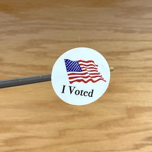 May include: A white round sticker with an American flag and the text "I Voted" in black.