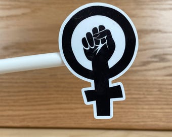 May include: A black and white sticker featuring the feminist symbol with a raised fist inside a circle. The symbol is a combination of the Venus symbol and a clenched fist, representing female empowerment and activism.