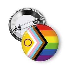 May include: A round button pin with a rainbow flag design and a second design with yellow, pink, blue, brown, and purple colors. The pin has a metal back with a safety pin clasp.