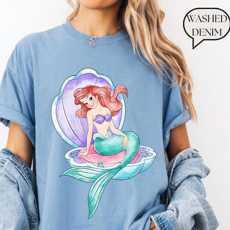 May include: Pale blue t-shirt with a mermaid graphic in a seashell. The mermaid has red hair, a purple seashell bra, and a teal tail. The text "Washed Denim" is in a speech bubble.