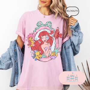 K&ouml;nnte beinhalten: Hellrosa T-Shirt mit einer Cartoon-Illustration von Arielle, der Meerjungfrau. Das Design umfasst Arielle, Fabius, Sebastian und Seepferdchen in einem dekorativen Rahmen. Das Shirt hat das Wort "Blossom" in einer Sprechblase.
