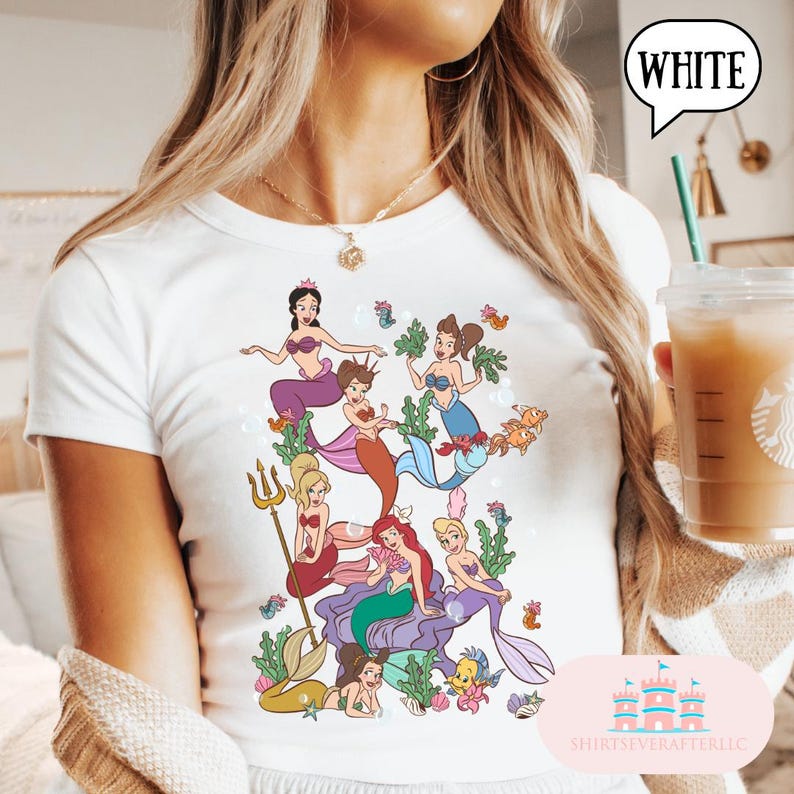 Op de afbeelding: Wit t-shirt met een kleurrijke illustratie van zeemeerminnen. Het ontwerp toont verschillende zeemeerminnen met diverse haarkleuren en outfits, omringd door zeedieren en bellen. Het woord "WHITE" staat in een tekstballon.