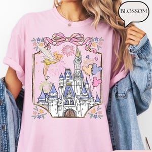 Puede incluir: Camiseta rosa claro con una ilustraci&oacute;n de castillo, un hada, fuegos artificiales y globos. El dise&ntilde;o est&aacute; enmarcado por un lazo rosa y detalles florales. La palabra "BLOSSOM" est&aacute; en una burbuja.