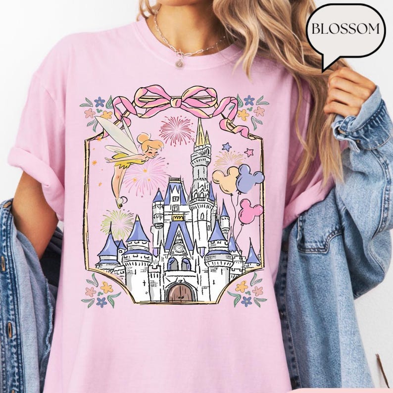 Puede incluir: Camiseta rosa claro con una ilustraci&oacute;n de castillo, un hada, fuegos artificiales y globos. El dise&ntilde;o est&aacute; enmarcado por un lazo rosa y detalles florales. La palabra "BLOSSOM" est&aacute; en una burbuja.