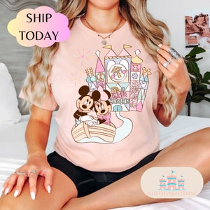 May include: Pink t-shirt with a graphic of Mickey and Minnie Mouse in a boat in front of a colorful castle with the text "It's a small world!"