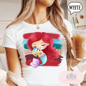 Puede incluir: Camiseta blanca con un dise&ntilde;o de dibujos animados de una sirena pelirroja abrazando a un pez azul y amarillo. El fondo presenta pinceladas rojas, azules y doradas. La palabra "WHITE" est&aacute; en un bocadillo.