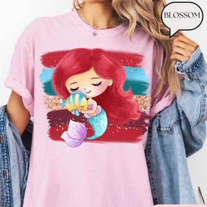 Puede incluir: Camiseta rosa claro con un dise&ntilde;o de sirena de dibujos animados. La sirena tiene el pelo rojo, una cola turquesa y sostiene un pez amarillo y azul. El dise&ntilde;o incluye pinceladas rojas, turquesas y doradas. La palabra "BLOSSOM" est&aacute; en una burbuja.
