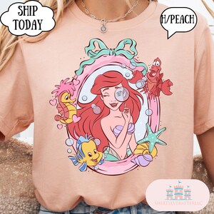 May include: Peach-colored t-shirt featuring a cartoon illustration of Ariel from The Little Mermaid. The design includes Ariel, Flounder, Sebastian, and a seahorse within a pink frame. The shirt has the text "Ship Today" and "H/Peach".