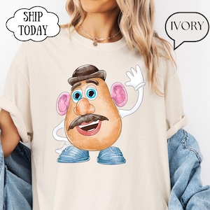 Può includere: Una t-shirt color avorio con un'illustrazione grafica di un personaggio a forma di patata dei cartoni animati con cappello marrone, baffi, orecchie rosa, scarpe blu e guanti bianchi. Il personaggio saluta con una mano. Ha un girocollo.