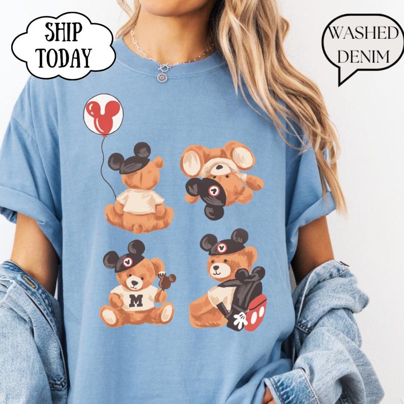 May include: A light blue t-shirt featuring a graphic print of four brown teddy bears wearing black mouse ear hats. One bear holds a red balloon, another wears a white shirt with an M, and one carries a backpack. The text reads SHIP TODAY and WASHED DENIM.