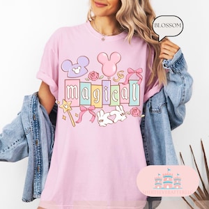 Op de afbeelding: Lichtroze t-shirt met het woord "magical" in een kleurrijk, cartoonachtig lettertype. Het ontwerp bevat Mickey Mouse oren, een toverstaf, strikken en rozen. Het shirt heeft een comfortabele, oversized pasvorm.