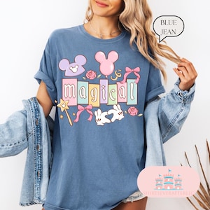 Op de afbeelding: Blauw spijkerbroekkleurig T-shirt met het woord "magical" in pastelkleuren, samen met cartoonafbeeldingen van een muis, konijn, ballonnen, strikken en rozen. Het shirt heeft opgerolde mouwen en een relaxte pasvorm.