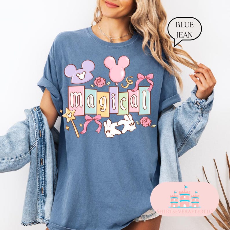 Op de afbeelding: Blauw spijkerbroekkleurig T-shirt met het woord "magical" in pastelkleuren, samen met cartoonafbeeldingen van een muis, konijn, ballonnen, strikken en rozen. Het shirt heeft opgerolde mouwen en een relaxte pasvorm.