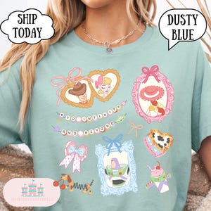 Op de afbeelding: Stofblauwe t-shirt met illustraties van stripfiguren in sierlijke frames. Het shirt bevat de zin "To Infinity and Beyond" en afbeeldingen van een cowboy, een cowgirl en een ruimteranger.