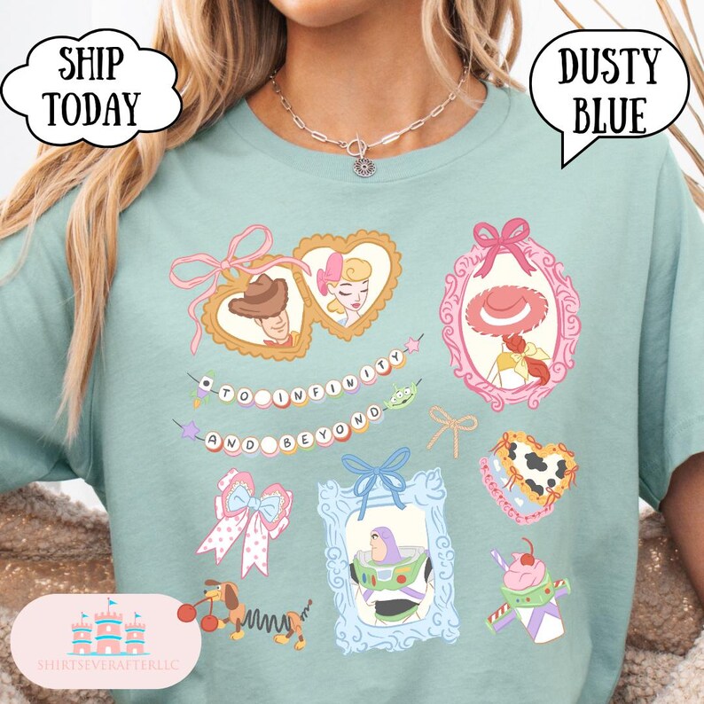 Op de afbeelding: Stofblauwe t-shirt met illustraties van stripfiguren in sierlijke frames. Het shirt bevat de zin "To Infinity and Beyond" en afbeeldingen van een cowboy, een cowgirl en een ruimteranger.