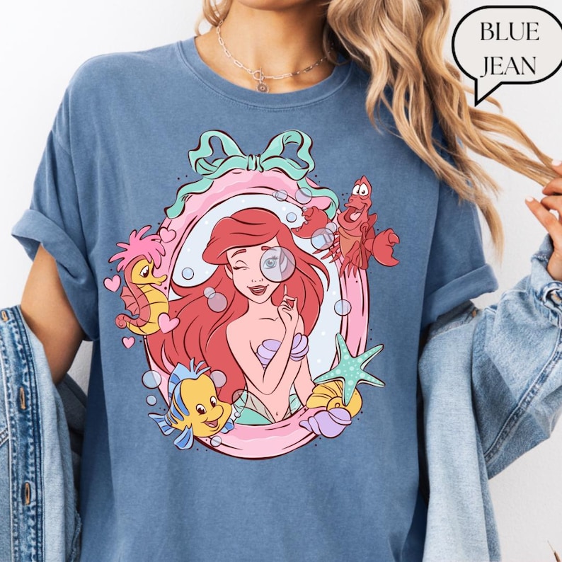 May include: Blue jean colored t-shirt featuring a cartoon illustration of Ariel from The Little Mermaid. The design includes Ariel, Flounder, Sebastian, and other sea creatures within a pink and green frame. The text "BLUE JEAN" is in a speech bubble.