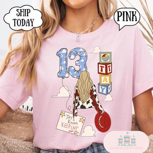 May include: A light pink t-shirt featuring a graphic of a person with long blonde hair wearing a cow-print jacket with red fringe. The design includes a blue cloud-patterned balloon shaped like the number 13, alphabet blocks, and a sign reading TAY'S ROOM KEEP OUT.