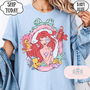 May include: Baby blue t-shirt featuring a cartoon illustration of a mermaid with red hair, a magnifying glass, and sea creatures. The design is framed by a pink and green border. Text on the shirt includes "SHIP TODAY" and "BABY BLUE".
