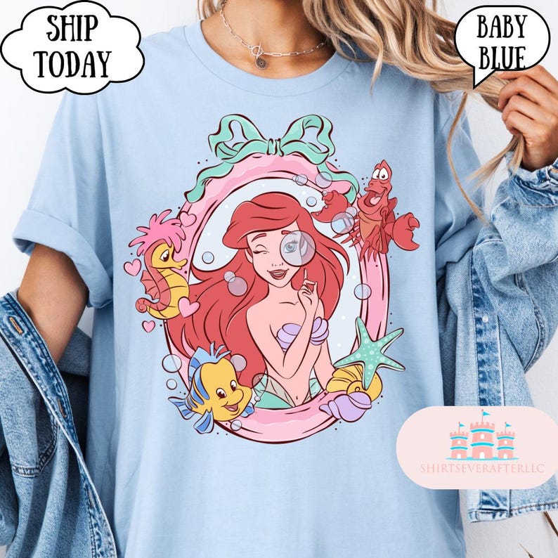 May include: Baby blue t-shirt featuring a cartoon illustration of a mermaid with red hair, a magnifying glass, and sea creatures. The design is framed by a pink and green border. Text on the shirt includes "SHIP TODAY" and "BABY BLUE".
