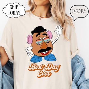 Può includere: Una t-shirt color avorio con la grafica di un personaggio a forma di patata marrone che indossa occhiali da sole con riflessi arcobaleno e castello. Il personaggio ha cappello nero, baffi, orecchie rosa e scarpe blu. Il testo dice Best Day Ever.